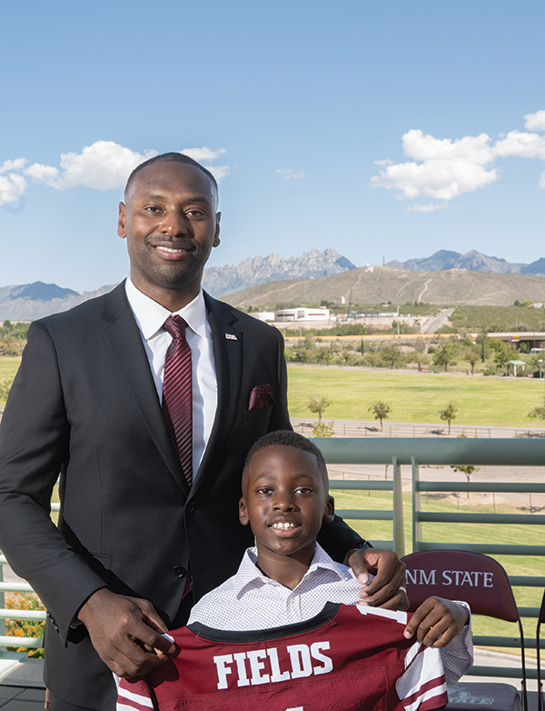 Man in a suit stands with a smiling boy holding a maroon sports jersey that reads “FIELDS,” with mountains and a blue sky in the background.