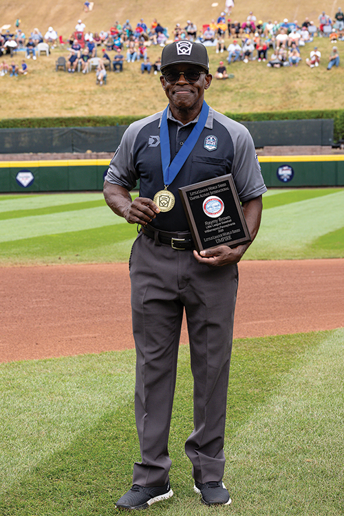 Baseball umpire on the field wearing sunglasses and a medal, holding an award plaque, with a crowd in the stadium seats behind him.