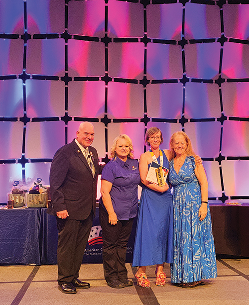 Four people pose on a stage at an awards event, with one person holding a plaque in front of a purple-and-pink lit backdrop.