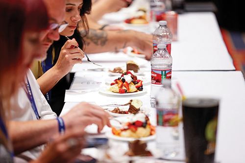 Several people seated at a long table sample plated desserts with fruit, with water bottles lined up along the table.